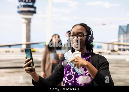 Heureuse voyageuse féminine dans l'aéroport prend selfies avec téléphone portable pour poster sur les médias sociaux pendant le voyage de vacances. Femme joyeuse tenant le billet d'avion prend des photos avec l'appareil photo de smartphone Banque D'Images