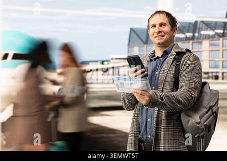 Portrait de l'homme souriant tenant smartphone et carte d'embarquement dans l'aéroport, faisant du sac à dos. Passager joyeux dans le terminal lumineux de l'aéroport à l'aide d'un téléphone portable Banque D'Images
