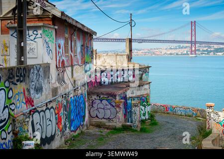LISBONNE, PORTUGAL - nov 09, 2025 : vieux bâtiments couverts de graffitis vibrants se dressent près du front de mer à Almada, Portugal, avec le 25 avril Bridg Banque D'Images