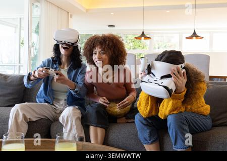 Famille féminine diversifiée assise sur un canapé dans le salon, deux portant des casques VR, une tenant du pop-corn Banque D'Images