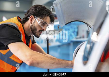 Technicien masculin portant un gilet de sécurité et des lunettes inspectant les composants des moteurs d'avion dans un hangar, représentant le service de l'industrie aéronautique, l'ingénierie Banque D'Images
