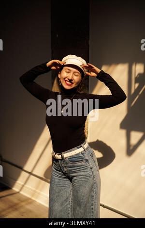 Une femme se tient dans une pièce avec une lumière vive et des ombres. Elle porte une casquette et une chemise noire à manches longues. Ses jeans sont larges et elle sourit en posant Banque D'Images