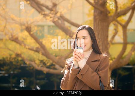 Heureuse femme japonaise profitant d'une pause café à l'extérieur parmi les arbres ginkgo d'automne Banque D'Images
