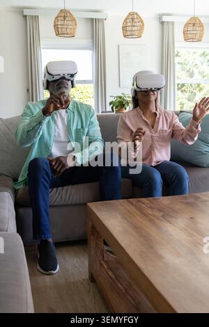 Couple senior diversifié assis sur un canapé gris dans le salon portant des casques blancs VR et atteignant Banque D'Images