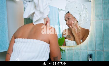 Femme noire appliquant des soins de la peau dans le miroir de salle de bain enveloppé dans la serviette après la douche avec la routine détendue Banque D'Images