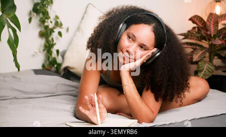 Femme noire se relaxant sur le lit avec des écouteurs journalisant et rêvant dans une chambre confortable Banque D'Images