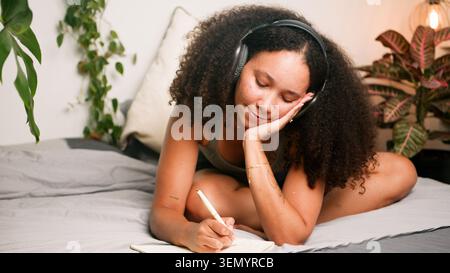 Femme noire se relaxant dans la chambre à coucher écrivant dans le journal tout en écoutant de la musique sur des écouteurs Banque D'Images