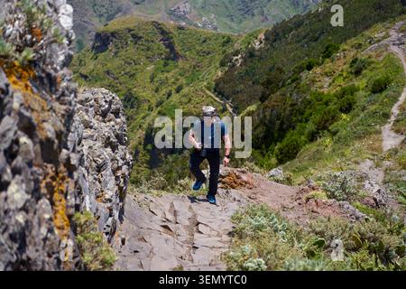 Photographe masculin avec appareil photo grimpant un sentier rocheux à marches sur une crête étroite dans les montagnes Anaga, Tenerife, avec des vallées verdoyantes en contrebas Banque D'Images