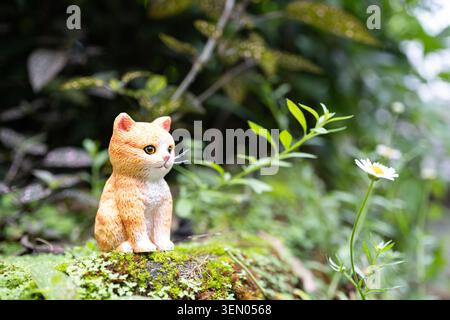Chiang mai, Thaïlande - 8 juin 2026 : figurine de chaton tabby orange assise sur la roche moussue à côté de fleurs sauvages de Marguerite dans un jardin tropical Banque D'Images