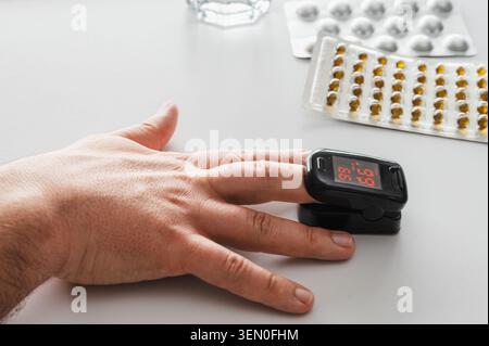 Gros plan d'un oxymètre de pouls du bout des doigts sur une main à côté d'un verre d'eau et de plaquettes thermoformées de pilules, illustrant la surveillance de la santé à domicile, l'oxygène du sang Banque D'Images