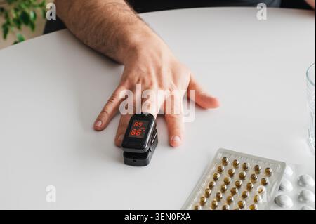 Gros plan d'un oxymètre de pouls du bout des doigts sur le doigt d'un homme mature à côté de plaquettes thermoformées de pilules, illustrant la surveillance de la santé à domicile, les paramètres vitaux vérifiant un Banque D'Images