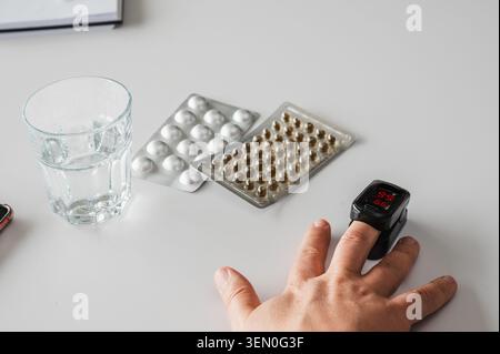 Gros plan d'un oxymètre de pouls du bout des doigts sur une main à côté d'un verre d'eau et de plaquettes thermoformées de pilules, illustrant la surveillance de la santé à domicile, l'oxygène du sang Banque D'Images