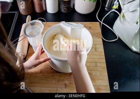 Vue aérienne des mains pliant la farine tamisée dans la pâte à gâteau éponge avec une spatule dans un bol dans une cuisine moderne, mélangeant la pâte pour un cak maison Banque D'Images