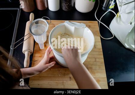 Vue aérienne des mains mélangeant la farine tamisée dans la pâte à gâteau éponge pâle dans un bol sur une planche de bois dans une cuisine moderne, préparant un gâteau fait maison. Banque D'Images