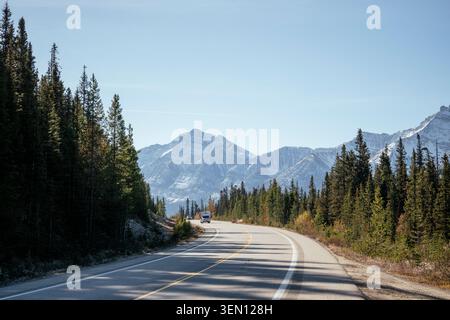 Une route sinueuse mène à travers une forêt avec des montagnes en arrière-plan sous un ciel bleu clair. Banque D'Images