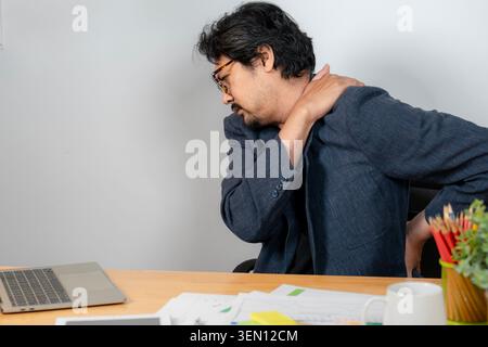 Un jeune homme d'affaires souffrant de douleurs au cou et à l'épaule alors qu'il travaillait à son bureau dans le bureau, représentant des symptômes du syndrome de bureau. Banque D'Images
