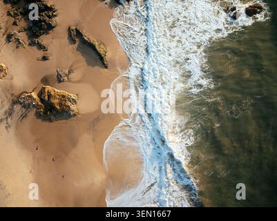 Vue aérienne des vagues qui s'écrasent sur une plage de sable rocheux pendant la journée. Banque D'Images