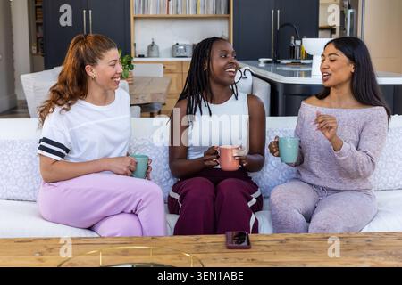 Diverses amies féminines assises sur le canapé dans le salon, tenant des tasses, smartphone reposant sur la table Banque D'Images