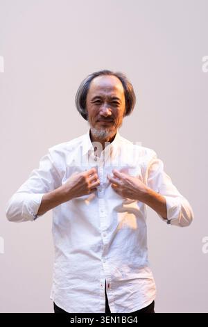 Ma Jian se tient dans un studio, souriant doucement pendant qu'il ajuste sa chemise. L'éclairage crée une atmosphère chaleureuse pendant qu'il se prépare à une interaction décontractée Banque D'Images