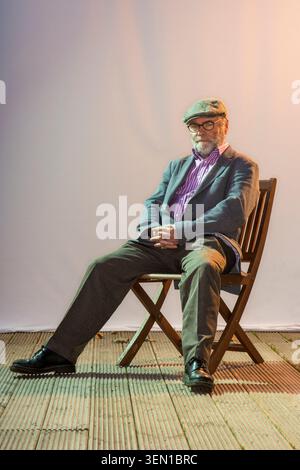 Malachi O'Doherty est assis sur une chaise en bois à l'intérieur. Il porte des lunettes et un bonnet plat tandis que la lumière crée une atmosphère chaleureuse derrière lui. Le spac Banque D'Images