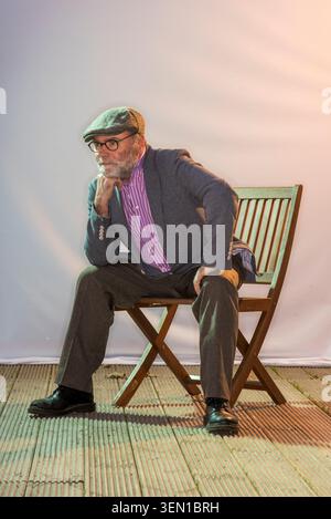 Malachi O'Doherty est assis sur une chaise en bois avec une main sur son menton. Il porte une casquette et une veste tout en paraissant réfléchi. Banque D'Images