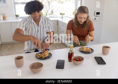 Couple en chemise rayée et haut de rouille riant, coupant des crêpes au bar du petit déjeuner avec une bouteille de sirop Banque D'Images