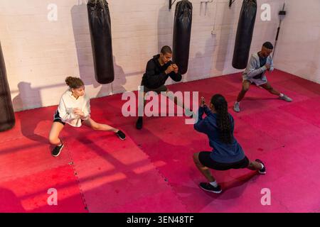 Groupe diversifié faisant des fentes latérales dans la salle de boxe, tapis rouges, sacs lourds avec adolescent, portant des vêtements de sport Banque D'Images