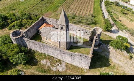 Vue de haut en bas par drone de l'église fortifiée de la Sainte Trinité à Hrastovlje, Slovénie, montrant les murs de pierre, le clocher, la cour, et des vignobles. Banque D'Images