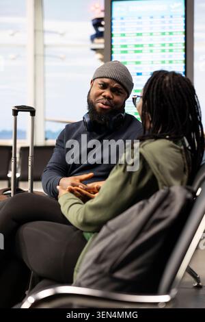 Des Noirs mariés se tenant la main et assis dans le hall de l'aéroport, attendant leur vol de lune de miel. Jeunes mariés planifiant un itinéraire touristique, souriant et appréciant le style de vie de loisirs. Banque D'Images