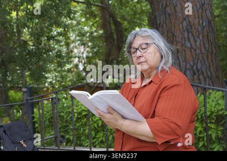 Femme senior profitant du temps calme, se concentrant sur un livre tout en étant assis sur un banc dans un environnement naturel de parc vert Banque D'Images