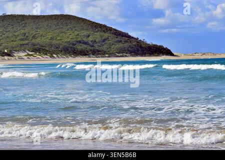 De douces vagues bleues se jettent sur une plage de sable pâle sous une haute colline densément boisée. Les nuages mous dérivent à travers des taches de ciel bleu, créant un calme, v Banque D'Images