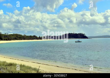 Une plage de sable rencontre une eau turquoise calme où un voilier repose près du rivage, soutenu par une forêt dense et verdoyante et une colline surmontée d'une petite tour. Banque D'Images
