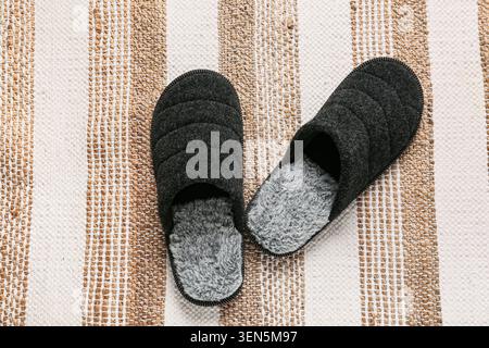 Chaussons de maison gris foncé confortables avec doublure en fausse fourrure douce sur un tapis tissé rayé, chaussures de maison confortables, les chaussures confortables offrent chaleur et Banque D'Images