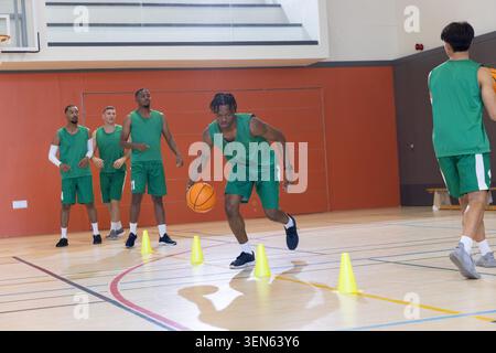 Divers coéquipiers masculins pratiquant l'exercice dribble dans le gymnase portant un équipement vert avec une balle orange, des cônes Banque D'Images