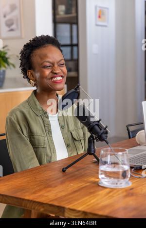 Afro-américaine souriante et enregistrement podcast à table avec micro à condensateur, ordinateur portable, verre Banque D'Images