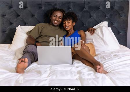 Couple afro-américain incliné dans le lit dans la chambre, portant un tee-shirt olive et un réservoir bleu, à l'aide d'un ordinateur portable Banque D'Images
