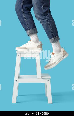 Personne en baskets blanches et jeans bleus montant un escabeau en bois blanc, isolé sur un fond bleu Banque D'Images
