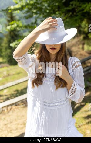Femme en robe blanche et chapeau de soleil. Portrait d'une femme portant une robe en dentelle blanche et un chapeau à large bord dans la lumière chaude du soleil près d'un chemin forestier. Banque D'Images