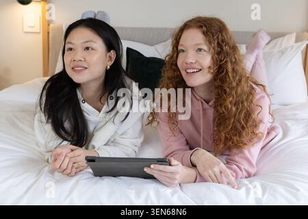 Deux amies allongées sur le lit dans la chambre appuyées sur les coudes portant des sweats à capuche partageant la tablette Banque D'Images