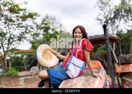 Femme chapeau tracteur ferme rurale chemise rouge sourire champ campagne, femme souriante tenant chapeau de paille assis sur le vieux tracteur dans la campagne agricole rurale avec Banque D'Images