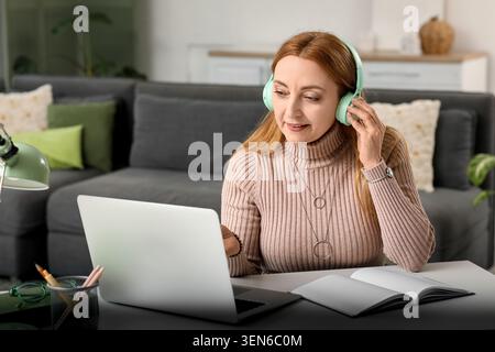 Femme mature dans des écouteurs travaillant à distance sur ordinateur portable à partir du bureau à domicile, l'apprentissage en ligne et le télétravail Banque D'Images