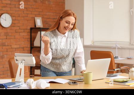 Femme d'affaires excitée célébrant le succès au bureau moderne avec ordinateur portable et tablette, atteignant des objectifs et la croissance financière Banque D'Images