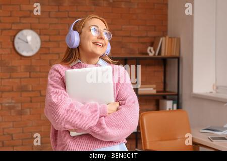 Femme souriante dans des écouteurs tenant un ordinateur portable, appréciant la musique ou podcast dans un environnement de bureau à domicile confortable, elle regarde vers le haut avec une expression joyeuse Banque D'Images