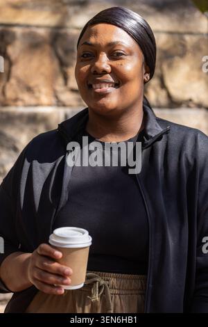 Femme afro-américaine souriante et tenant une tasse de café brune portant une veste noire contre un mur de pierre Banque D'Images