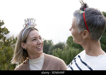 Femme dans les années 30 et femme senior debout dans le jardin portant bandeau de Noël et tiare rouge souriant Banque D'Images