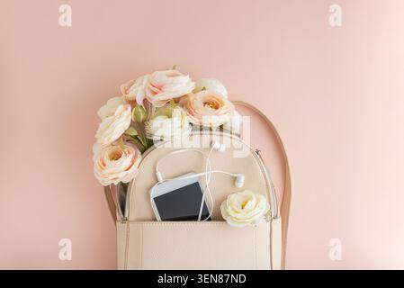 Sac à dos en simili cuir beige affichant un bouquet de fleurs rose pastel, un smartphone blanc et des écouteurs, créant une élégance fraîche et féminine et sp Banque D'Images