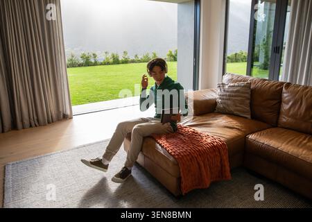 Homme asiatique assis sur une chaise brune dans le salon, portant une tablette à capuche et fixant le casque Banque D'Images