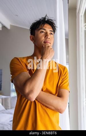 Homme asiatique debout par une grande fenêtre avec un rideau transparent dans la chambre, touchant le menton portant un t-shirt moutarde Banque D'Images