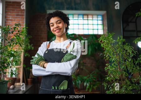 Gestion de sa propre entreprise. Femme africaine fleuriste portant tablier dans le magasin botanique avec des plantes vertes. Heureux propriétaire de petite entreprise travaillant au magasin de fleurs souriant entouré de petites entreprises de plantes Banque D'Images