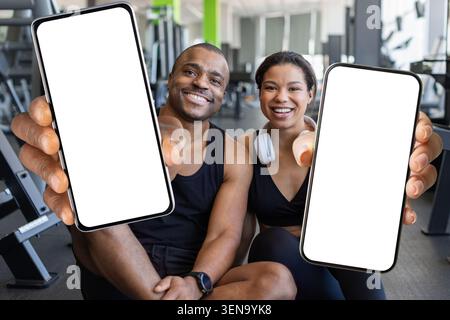 Jeune fitness noir assis dans le gymnase, tenant des smartphones avec des écrans vierges Banque D'Images